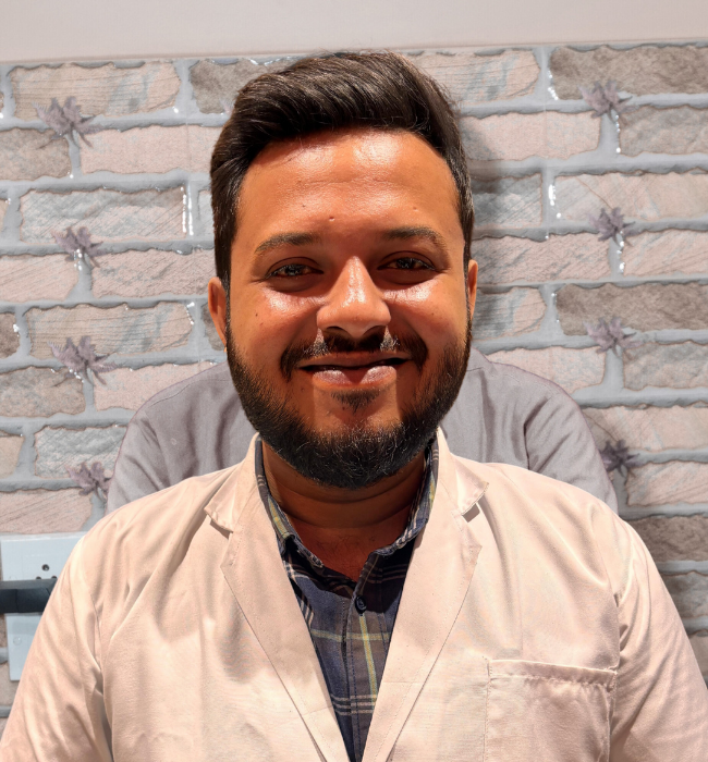 Male dental professional in a light lab coat smiling confidently against a brick-patterned clinic wall.
