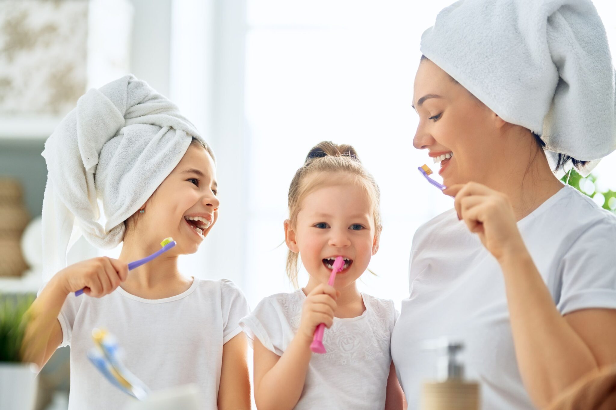 Mother and two young daughters brushing teeth together in bathroom, smiling and holding colorful toothbrushes.