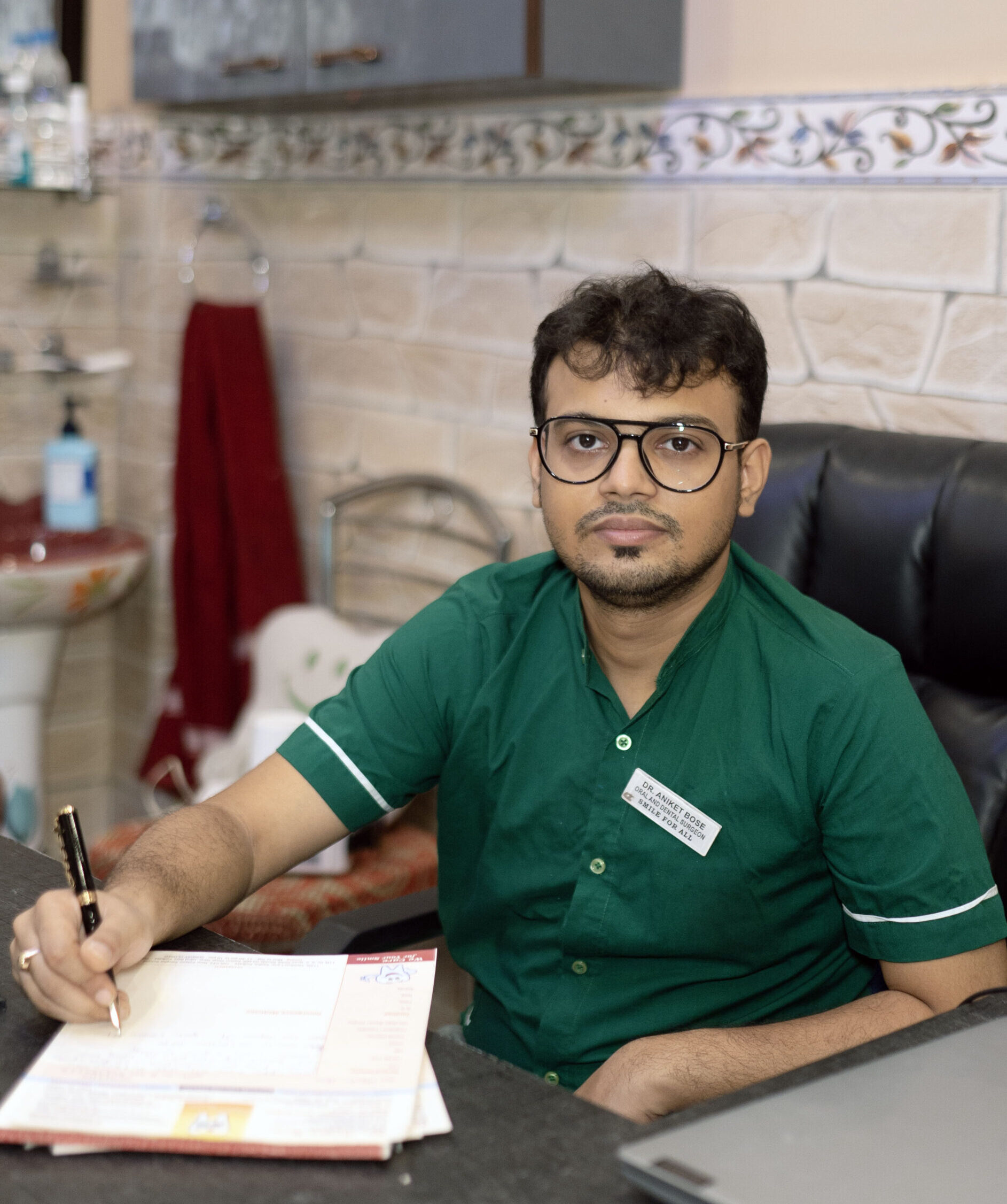 Dental professional in a green uniform seated at a clinic desk, reviewing patient documents in a warm, well-lit office.