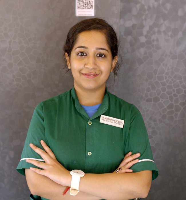 Female dental professional in a green uniform standing confidently with folded arms against a modern clinic background.