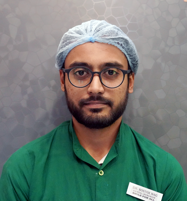 Male dental professional in a green uniform and surgical cap posing confidently against a modern clinic background.